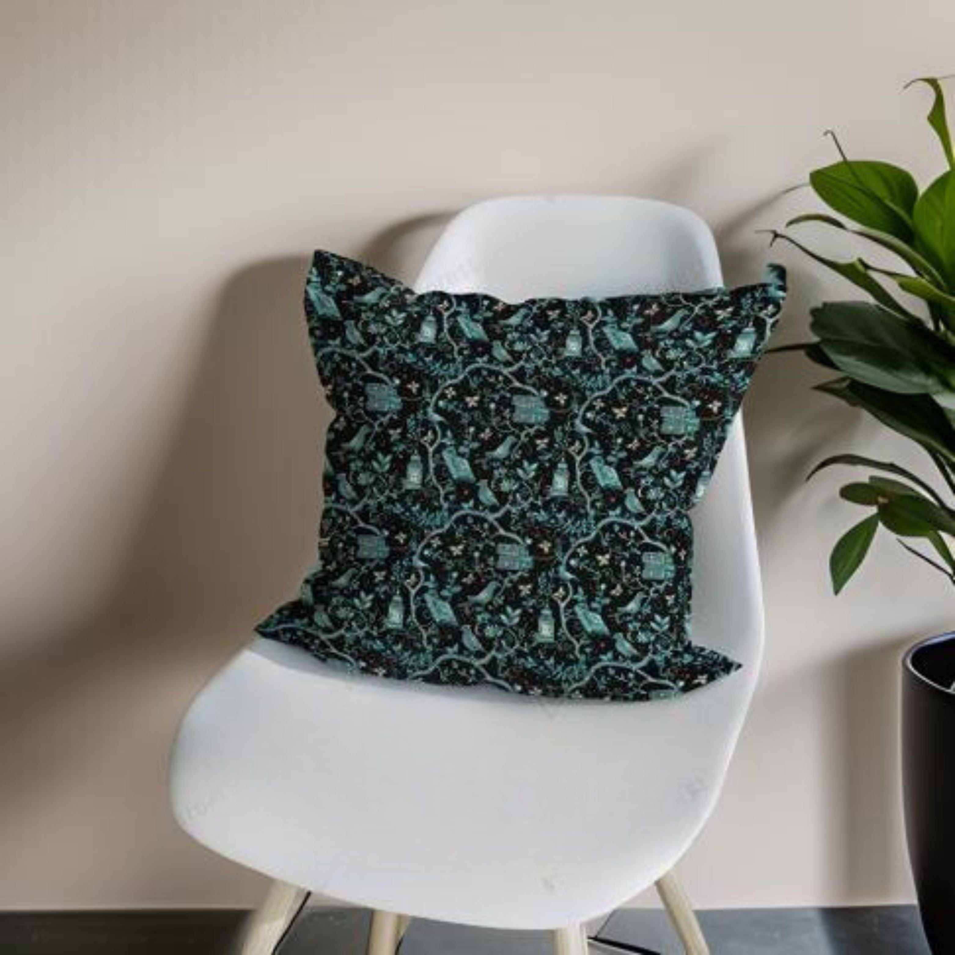 A square faux suede throw pillow with a whimsical pattern featuring teal birds and gothic motifs, placed on a white armchair against a pink wall with a green potted plant beside it.