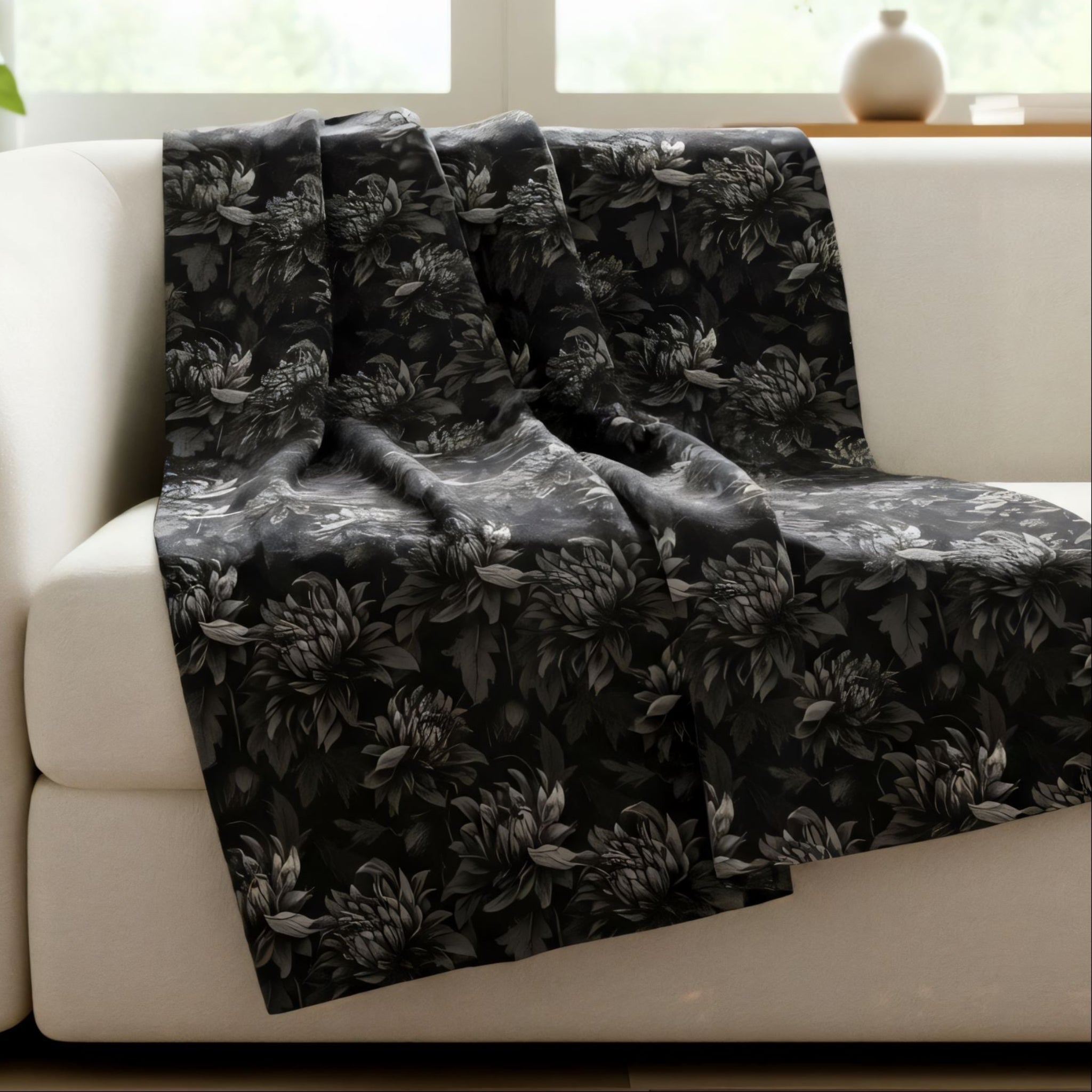 Black floral oversized blanket draped over a white sofa in a bright room with a plant and vase in the background.