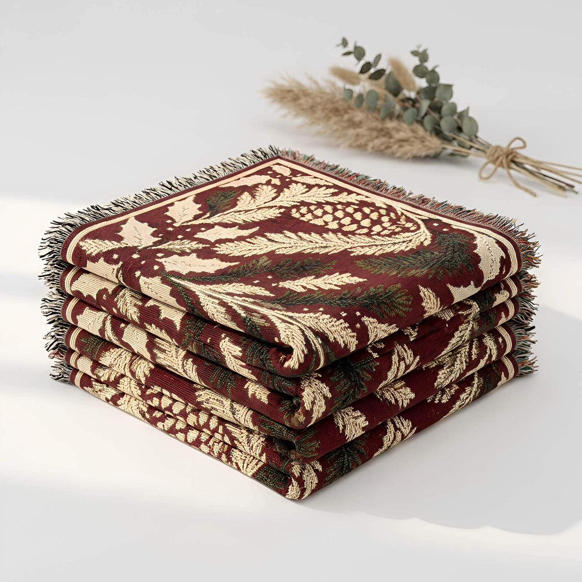 Folded Christmas woven cotton blanket showing its layered fringe and cranberry red botanical pattern with pinecones, holly, berries, and evergreen foliage.