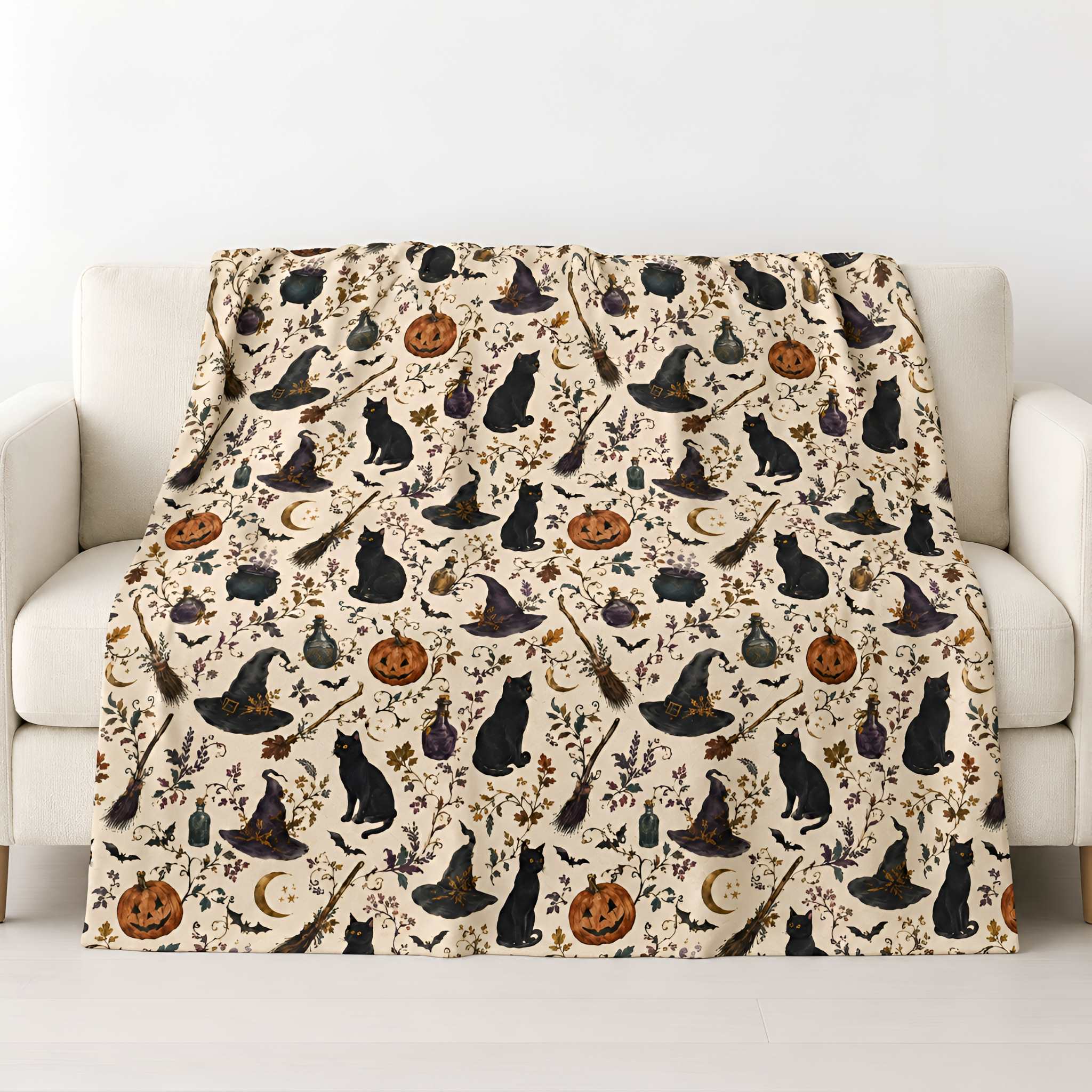 A cream-colored throw blanket with a repeating Halloween pattern of black cats, witch hats, broomsticks, pumpkins, and bats, draped over a white armchair.