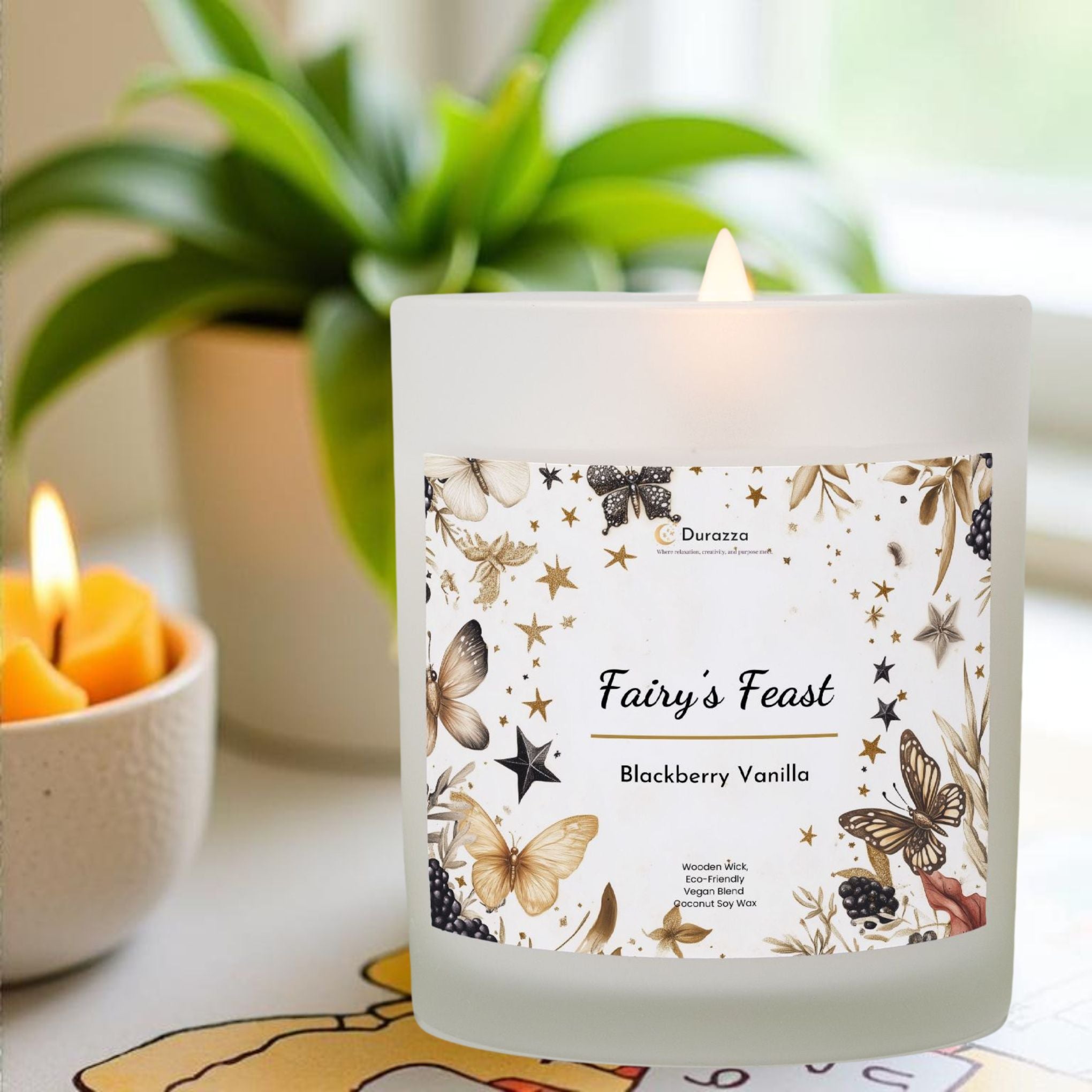 A lit Blackberry Vanilla scented candle labeled 'Fairy's Feast' with a wooden wick, placed on a table with a plant in the background.