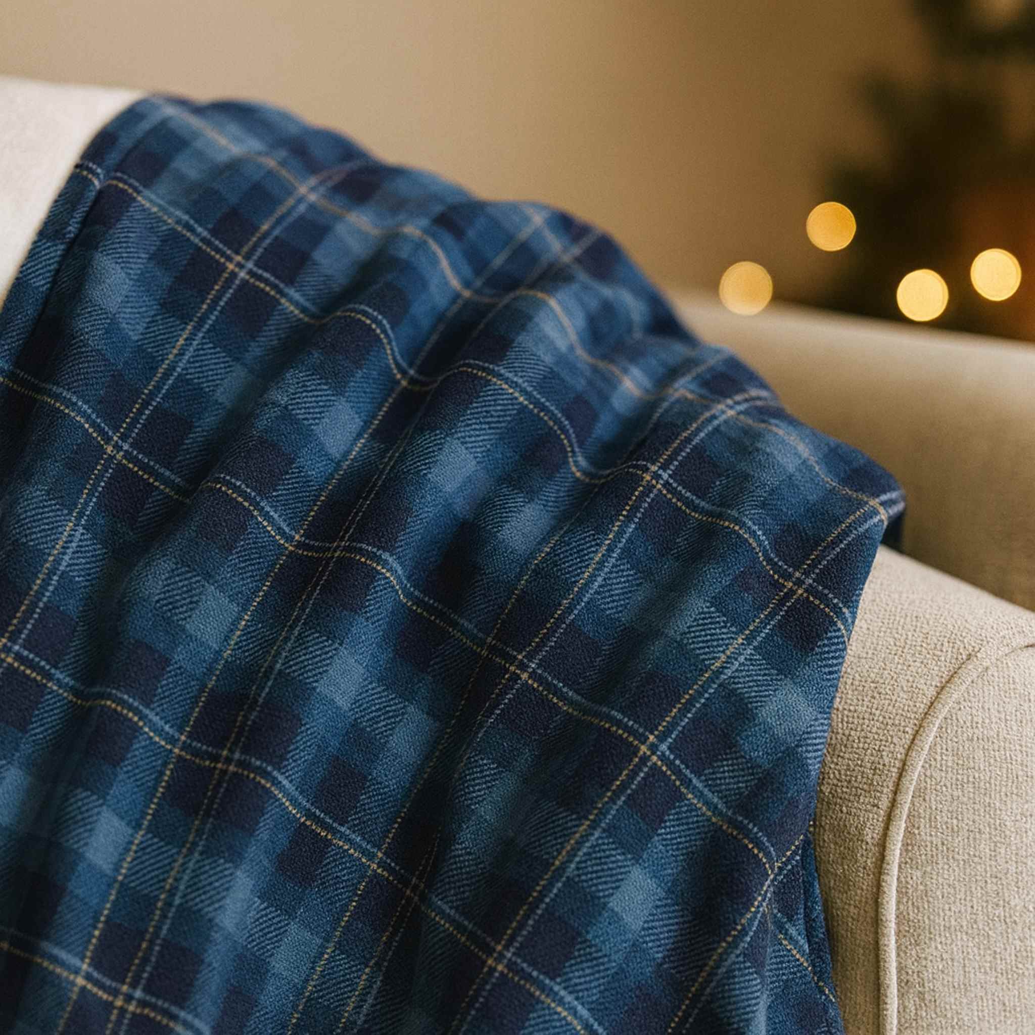 Blue plaid velveteen blanket draped over a beige couch with warm lights in the background