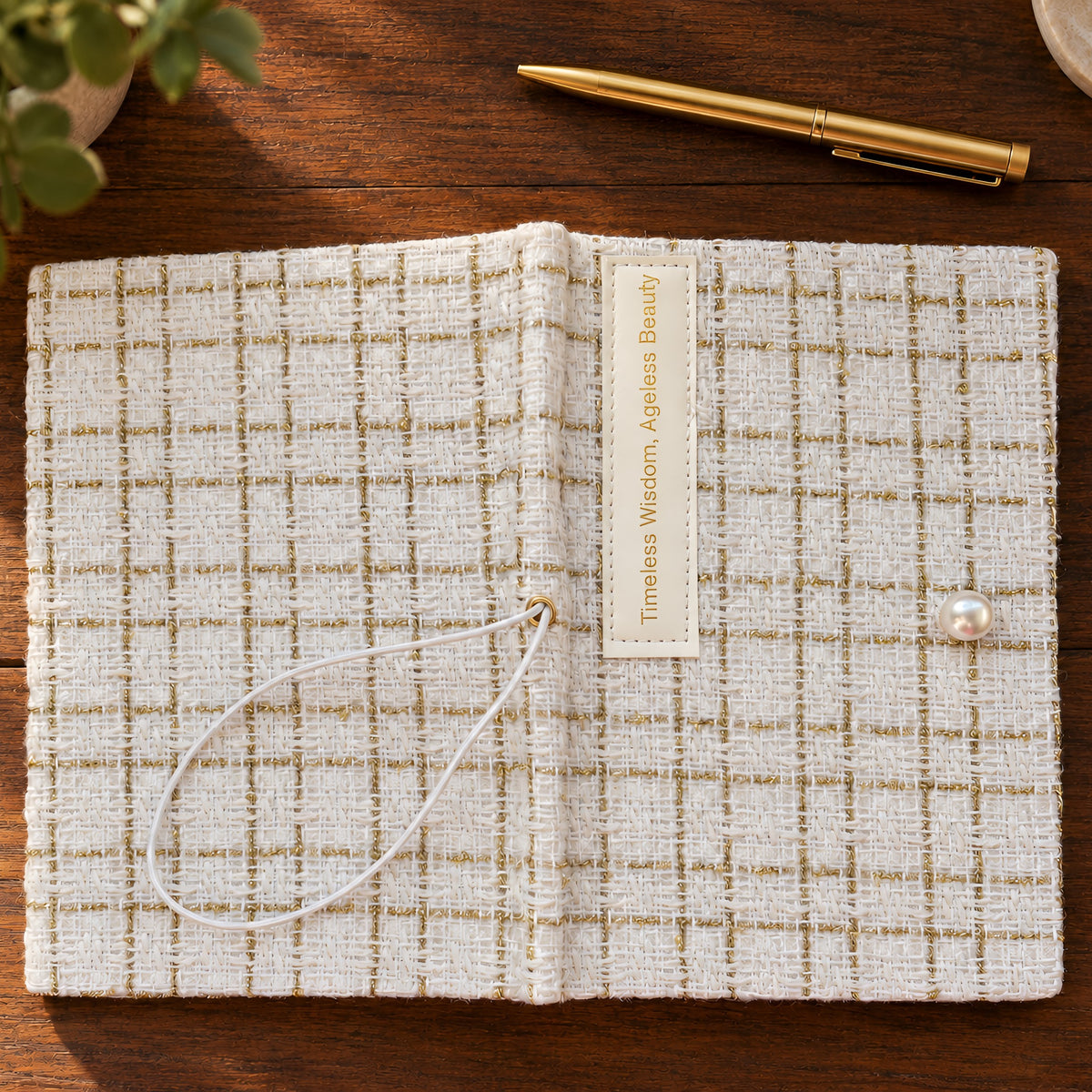 Cream fabric journal from above, showing gold-thread check, Timeless Wisdom label, and pearl-like accent on a wooden desk.
