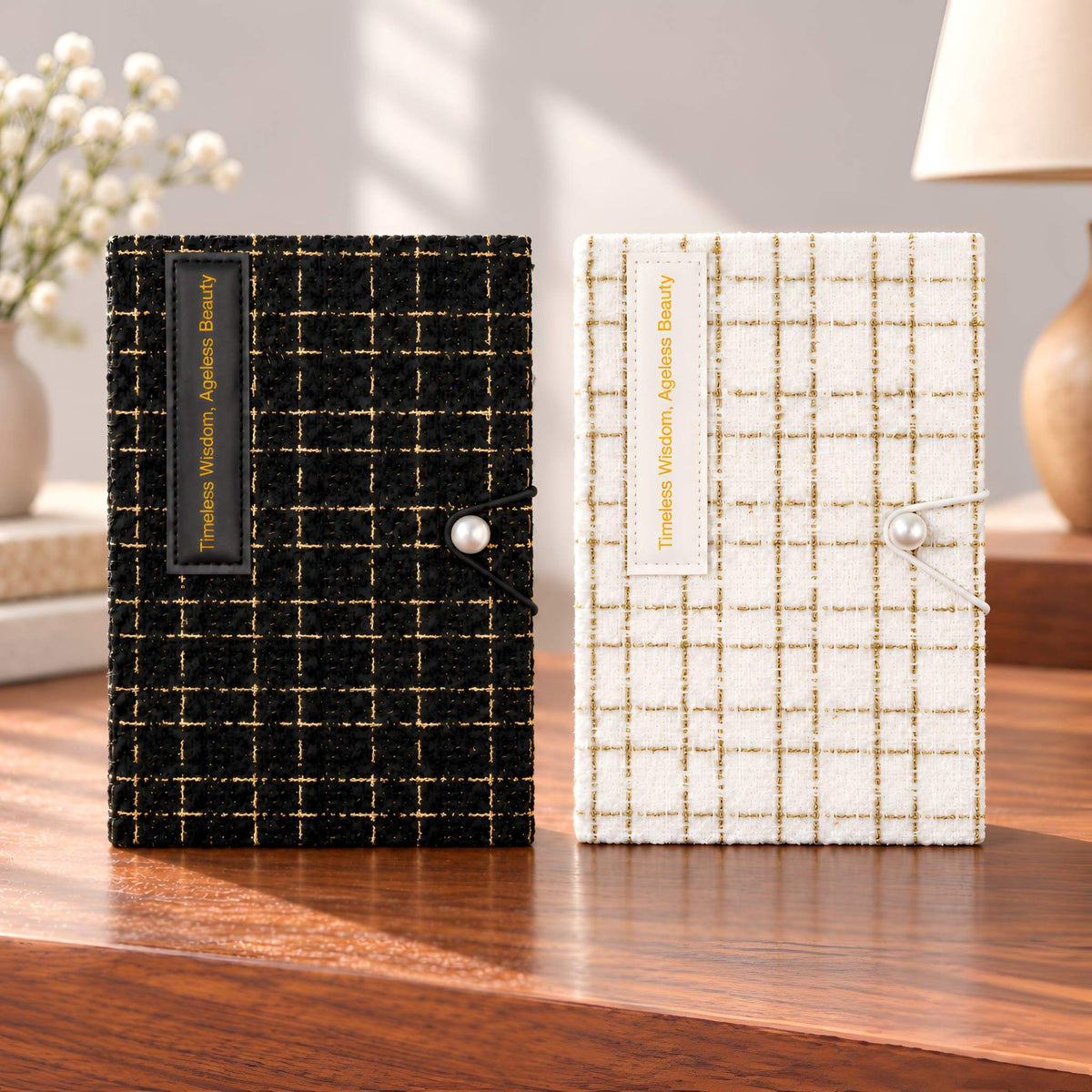 Black and cream fabric journals with gold-thread check covers and pearl-like accents displayed side by side on a wooden table.