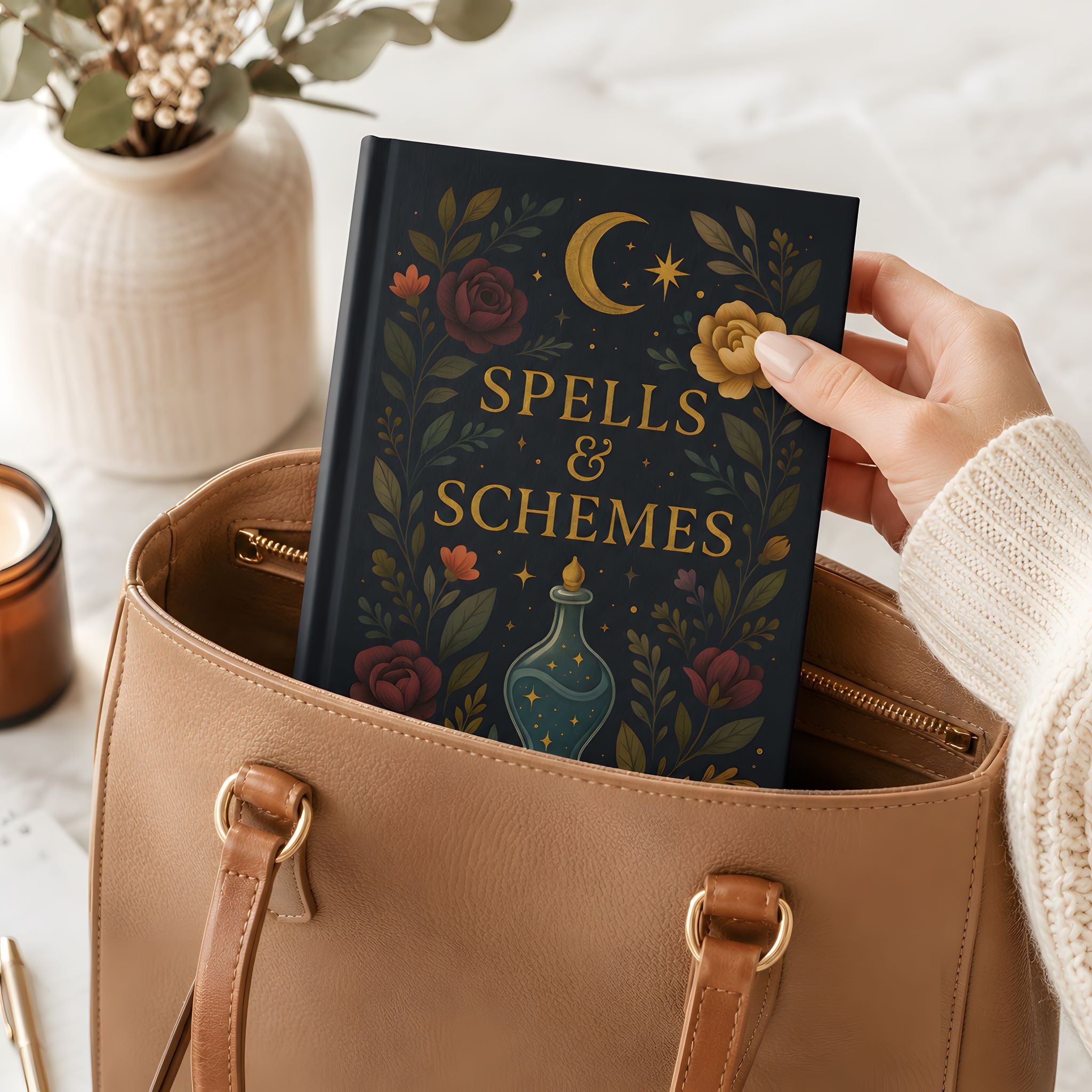 Whimsigoth hardcover journal titled 'Spells & Schemes' held by a hand over a beige handbag with decorative elements.