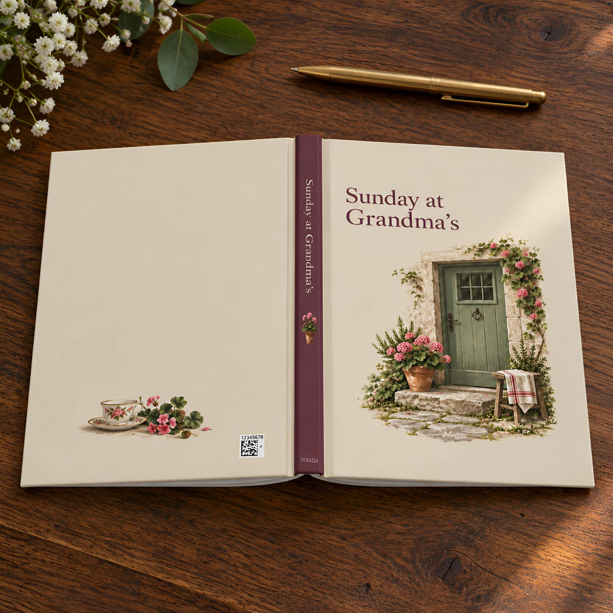 An open hardcover lined journal titled "Sunday at Grandma's" lies on a wooden tabletop, styled with a gold pen, small white flowers, and green leaves. The cover is cream with a burgundy spine and a printed illustration of a green door, flowers, and a small table setting.
