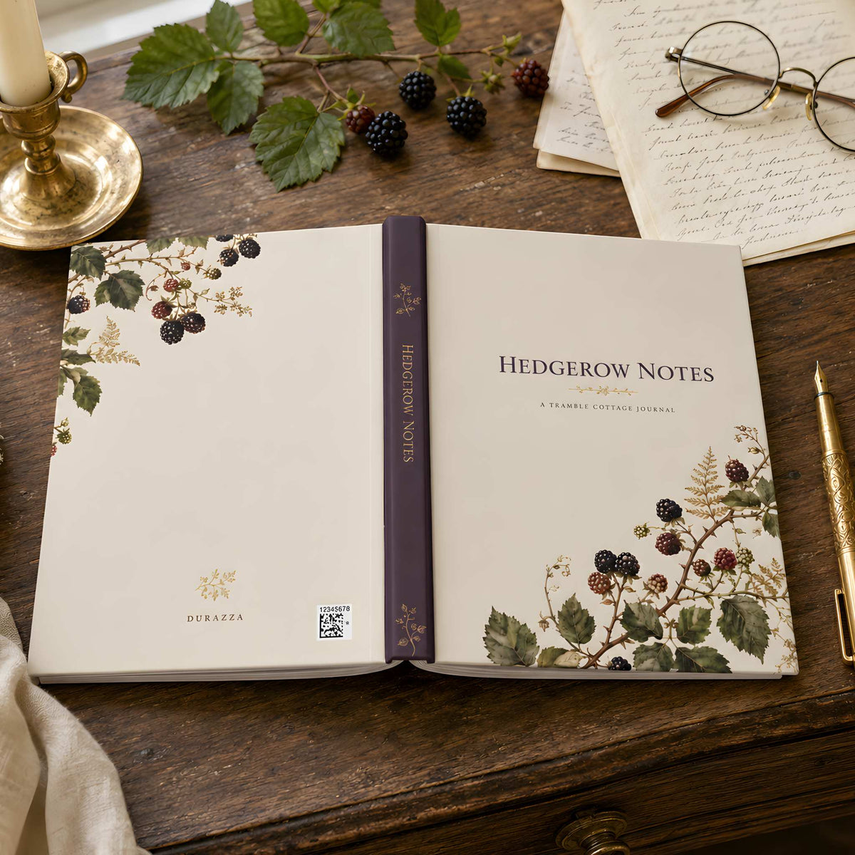 Open cream botanical journal showing its front, plum spine, and back blackberry artwork on a wooden desk with berries, candle, glasses, papers, and a gold pen.