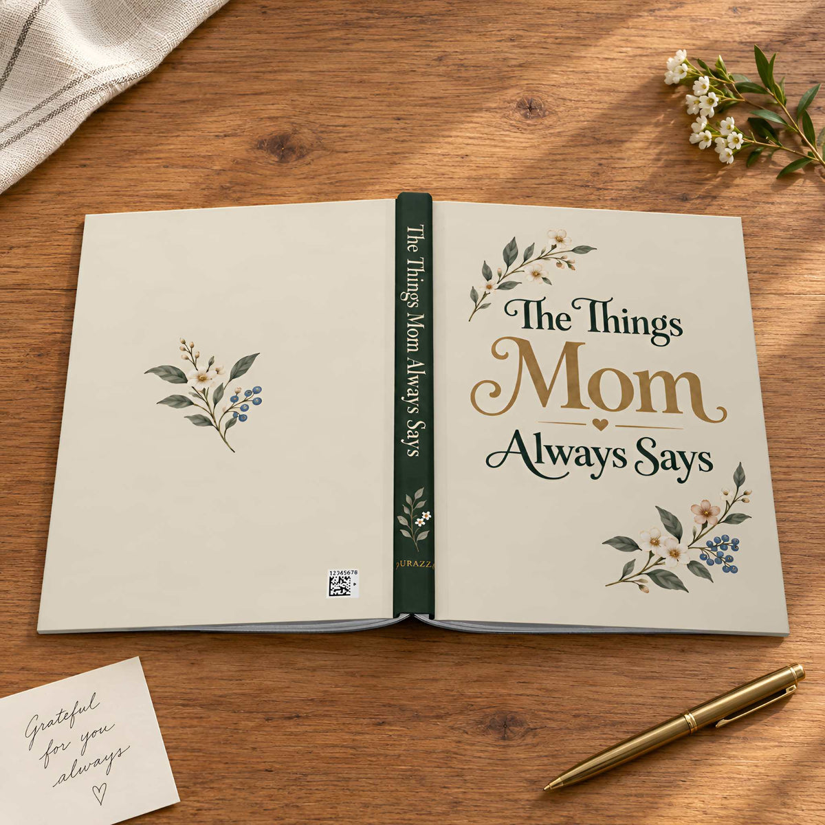 An open decorative journal on a wooden tabletop, titled "The Things Mom Always Says," with floral illustrations and elegant typography. A small note card and gold pen sit nearby, with a folded cloth and flowers in the background.