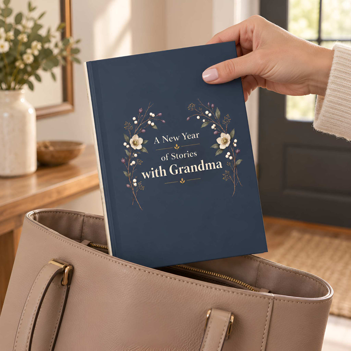 A navy blue hardcover journal with floral artwork and the text 'A New Year of Stories with Grandma' is being held in a person's hand and placed into a beige tote bag in a warm indoor setting.