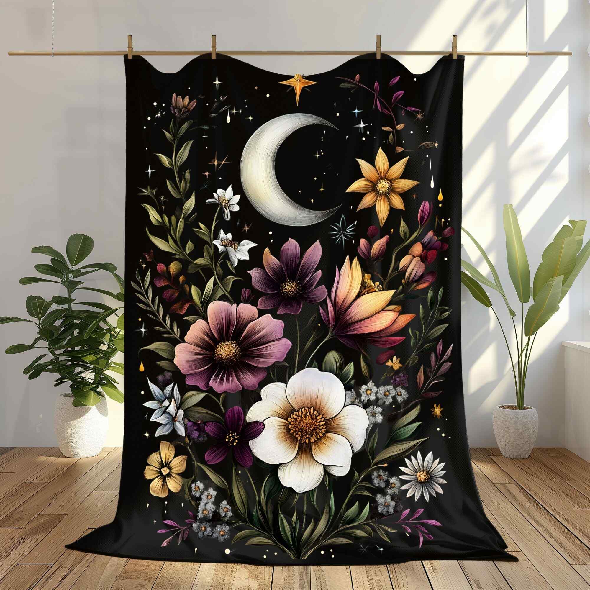 A black throw blanket with a floral and celestial design, featuring a variety of flowers, stars, and a crescent moon, hung on a wooden rod with a plant in the background.