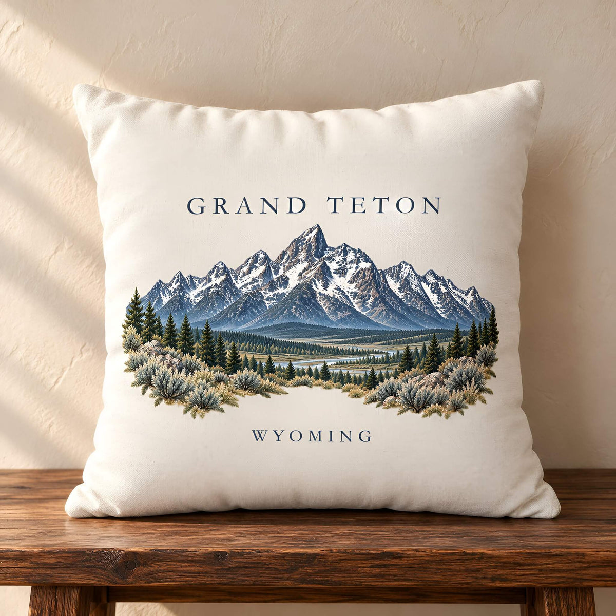 A square off-white throw pillow cover featuring a printed illustration of the Grand Teton mountains in Wyoming, resting on a dark wooden table against a textured beige wall.