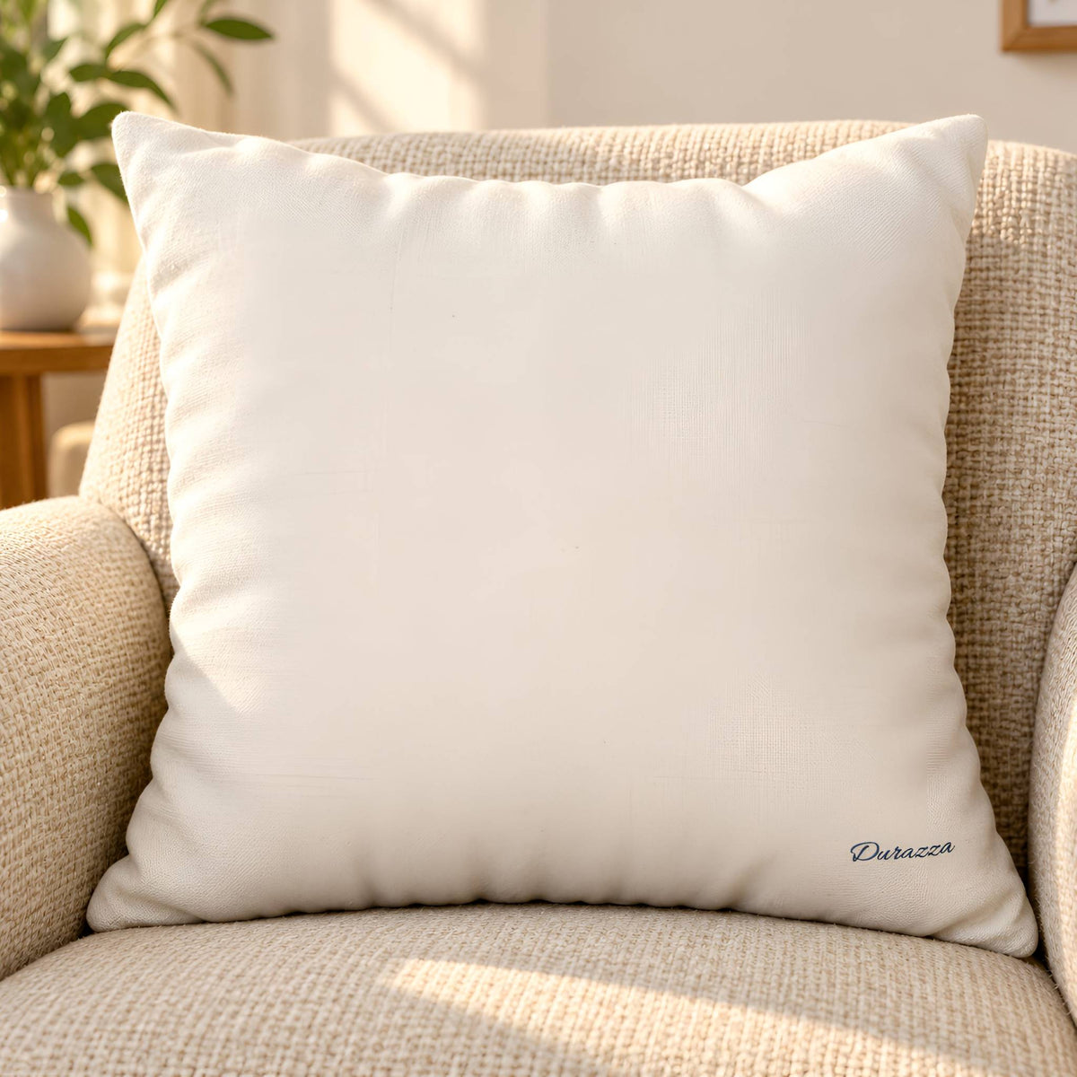A square backside of an off-white throw pillow with the name 'Durazza' printed in navy blue script in the bottom right corner, resting on a textured beige armchair.