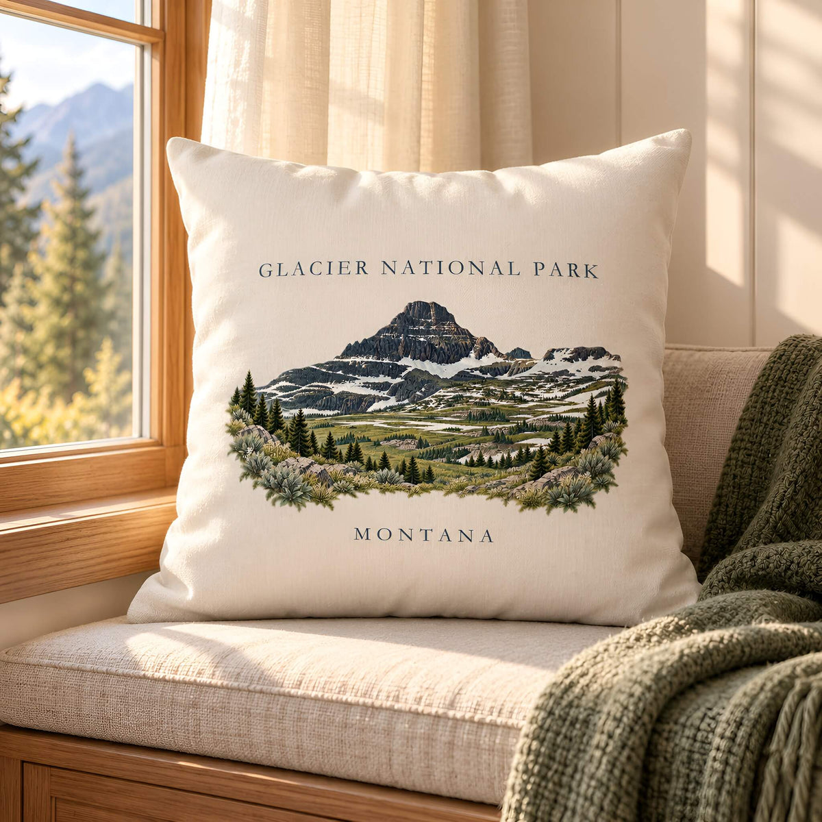 A square cream throw pillow featuring an illustrated Glacier National Park mountain scene with the text 'GLACIER NATIONAL PARK' and 'MONTANA', styled on a cushioned window seat with a window, curtains, and a green knit blanket nearby.