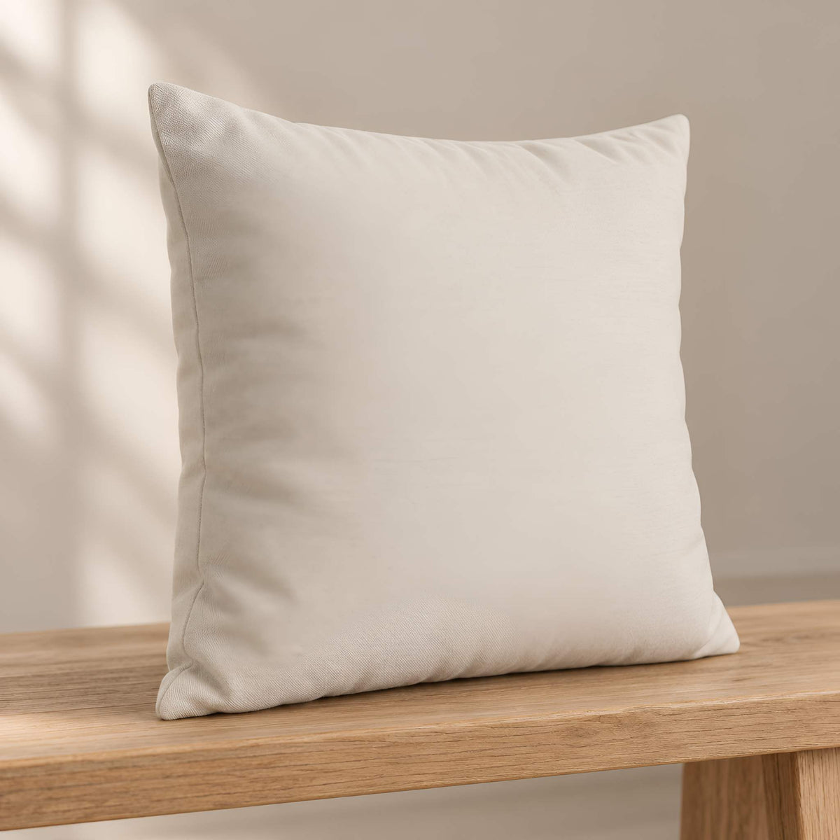 A square off-white throw pillow cover with a soft, textured fabric, resting on a light wood table against a neutral beige wall with soft window shadow patterns.