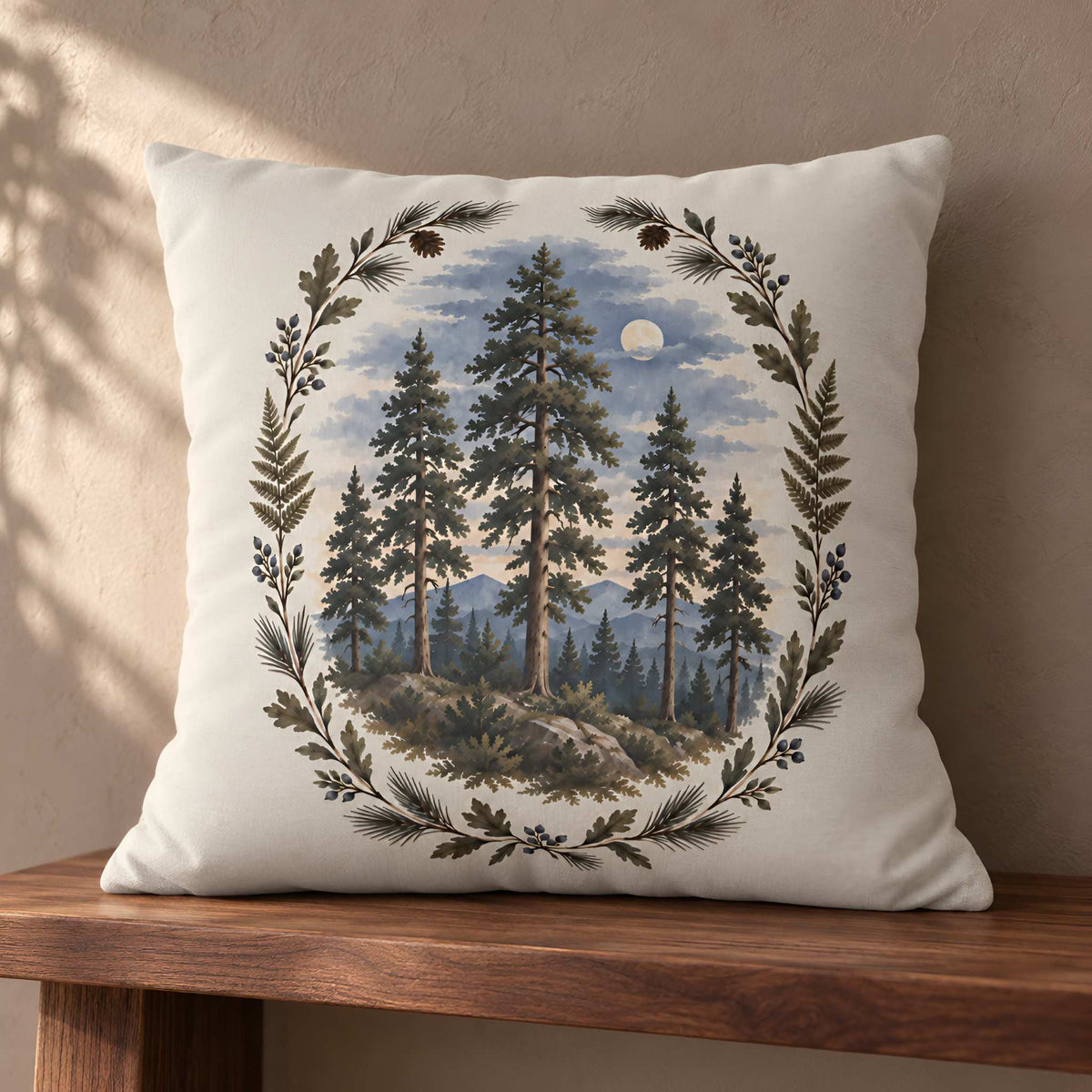 A square cream throw pillow cover with a detailed forest landscape print, featuring pine trees, mountains, and a moon surrounded by a leafy wreath, resting on a wooden bench against a textured beige wall.