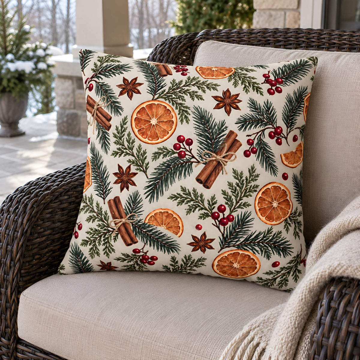 A square decorative throw pillow cover with a festive holiday print featuring pine branches, cinnamon sticks, star anise, red berries, and orange slices on an off-white background, styled on a wicker armchair with a beige cushion and throw blanket.