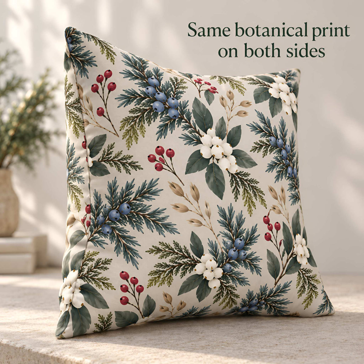 A square decorative throw pillow with a botanical print of green foliage, white flowers, and red berries on a cream background, styled on a stone surface with text overlay reading 'Same botanical print on both sides'.