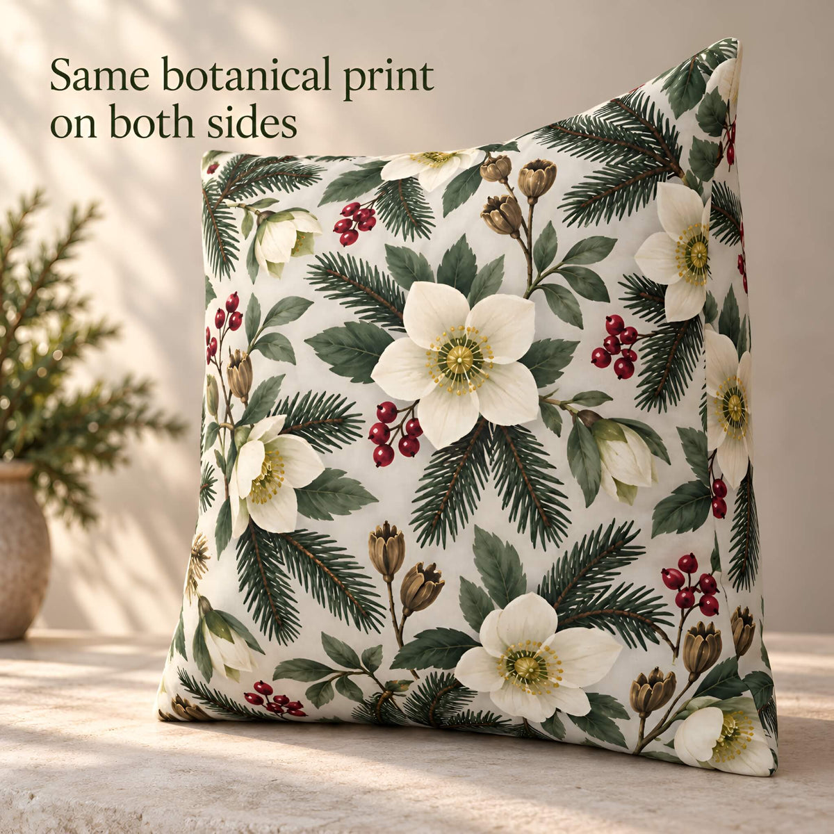 A square decorative throw pillow with a botanical print of white flowers, green pine needles, and red berries on a cream background, styled on a stone surface with a potted plant in the background. Text in the upper left reads 'Same botanical print on both sides'.