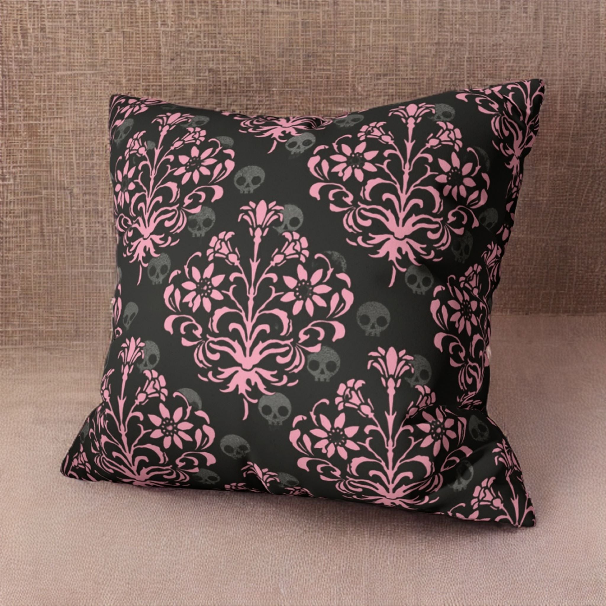 Decorative pillow with a black base and pink floral pattern featuring white skulls, presented on a textured background.