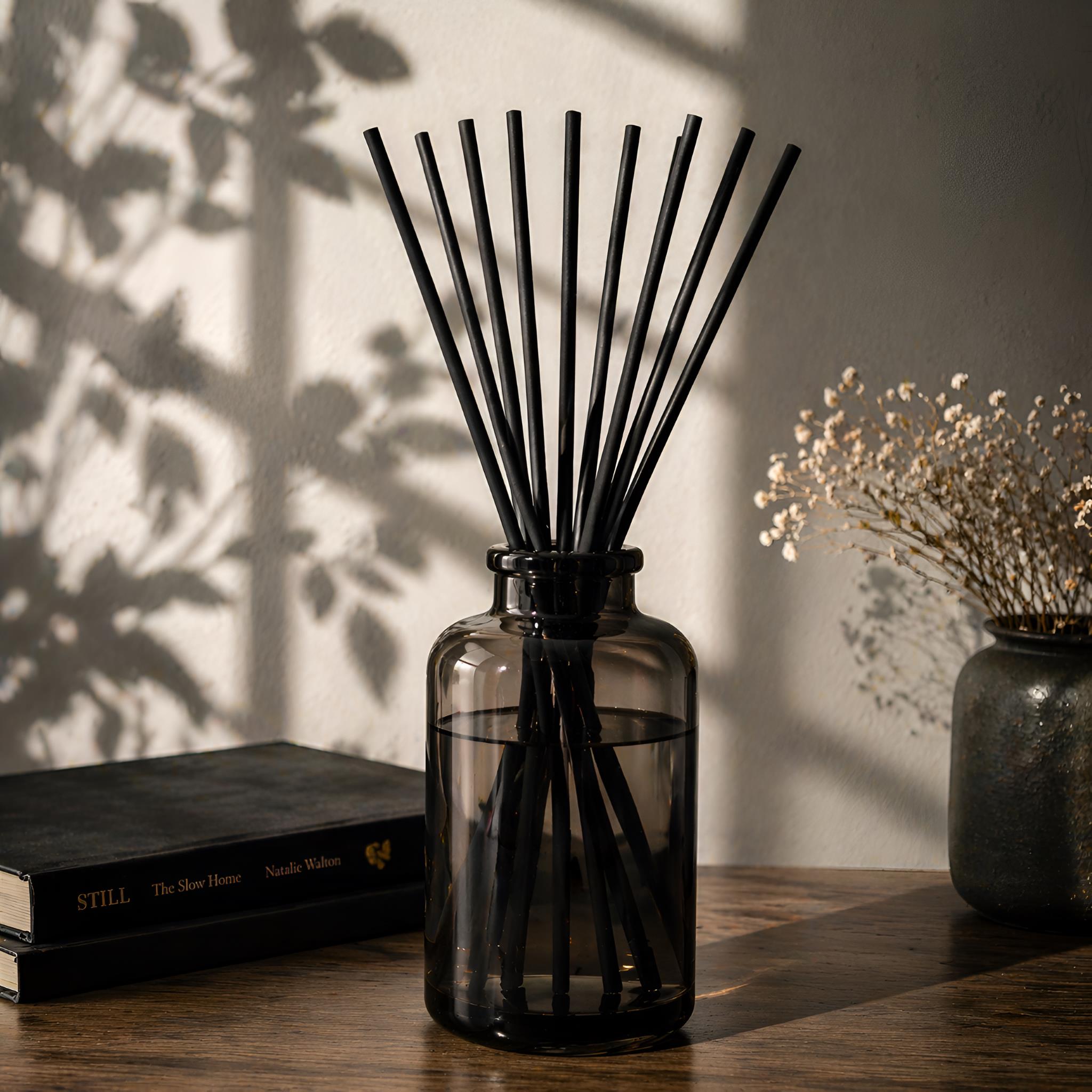 Black reed diffuser in a glass bottle on a wooden surface with books and dried flowers in the background.