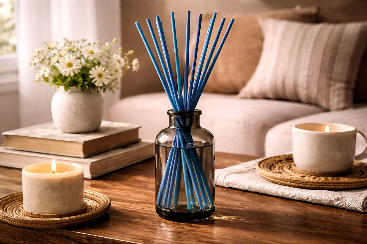 Blue diffuser reeds in a tall jar with a candle, books, flowers on a wooden table.
