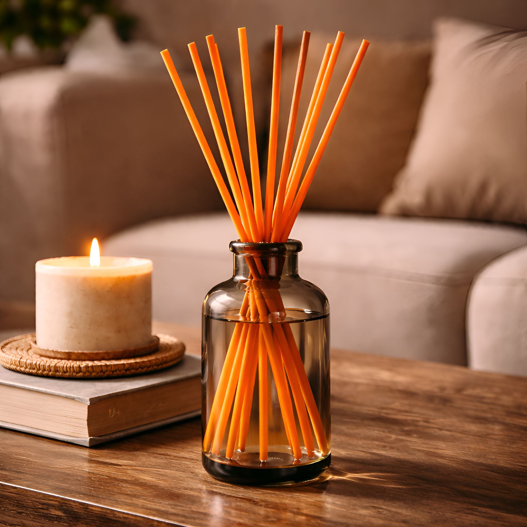 Orange Reed diffuser sticks in a tall smoky, clear glass bottle on a wooden table with a candle and books in the background.