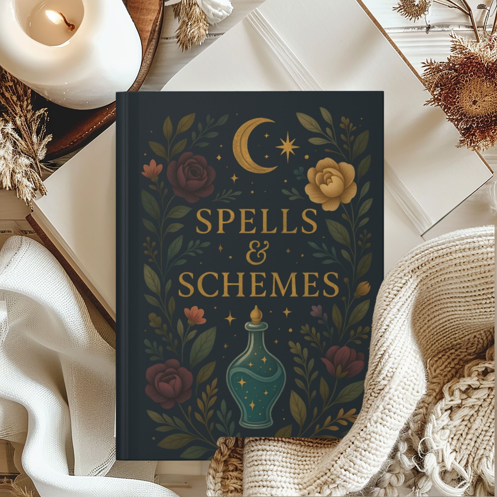 Journal titled 'Spells & Schemes' with decorative cover featuring flowers and a moon, placed on a surface with candles and plants.