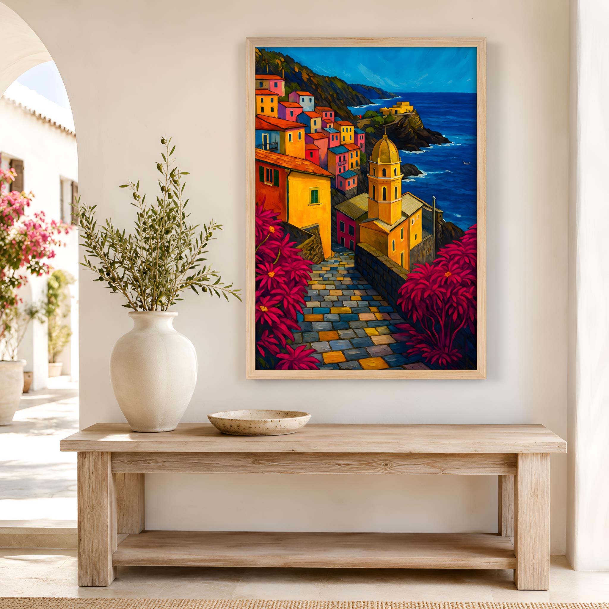 Framed colorful Mediterranean painting of a coastal village on a wall above a wooden bench with a vase and flowers.