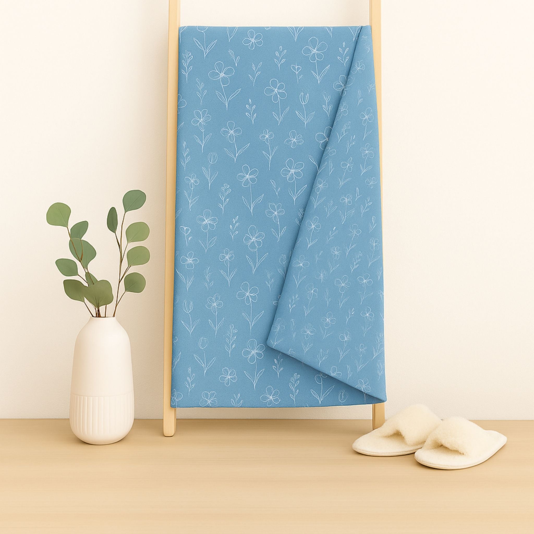 Blue throw blanket with a white floral pattern draped over a wooden ladder, next to a white vase with greenery on a beige surface.