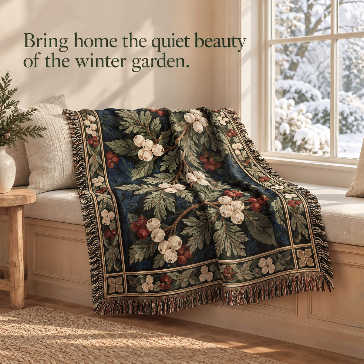 A decorative cotton throw blanket with a winter garden floral pattern is draped over a wooden window seat in a sunlit room. The blanket features cream blossoms, green leaves, and red berries on a dark navy background with a fringed edge.