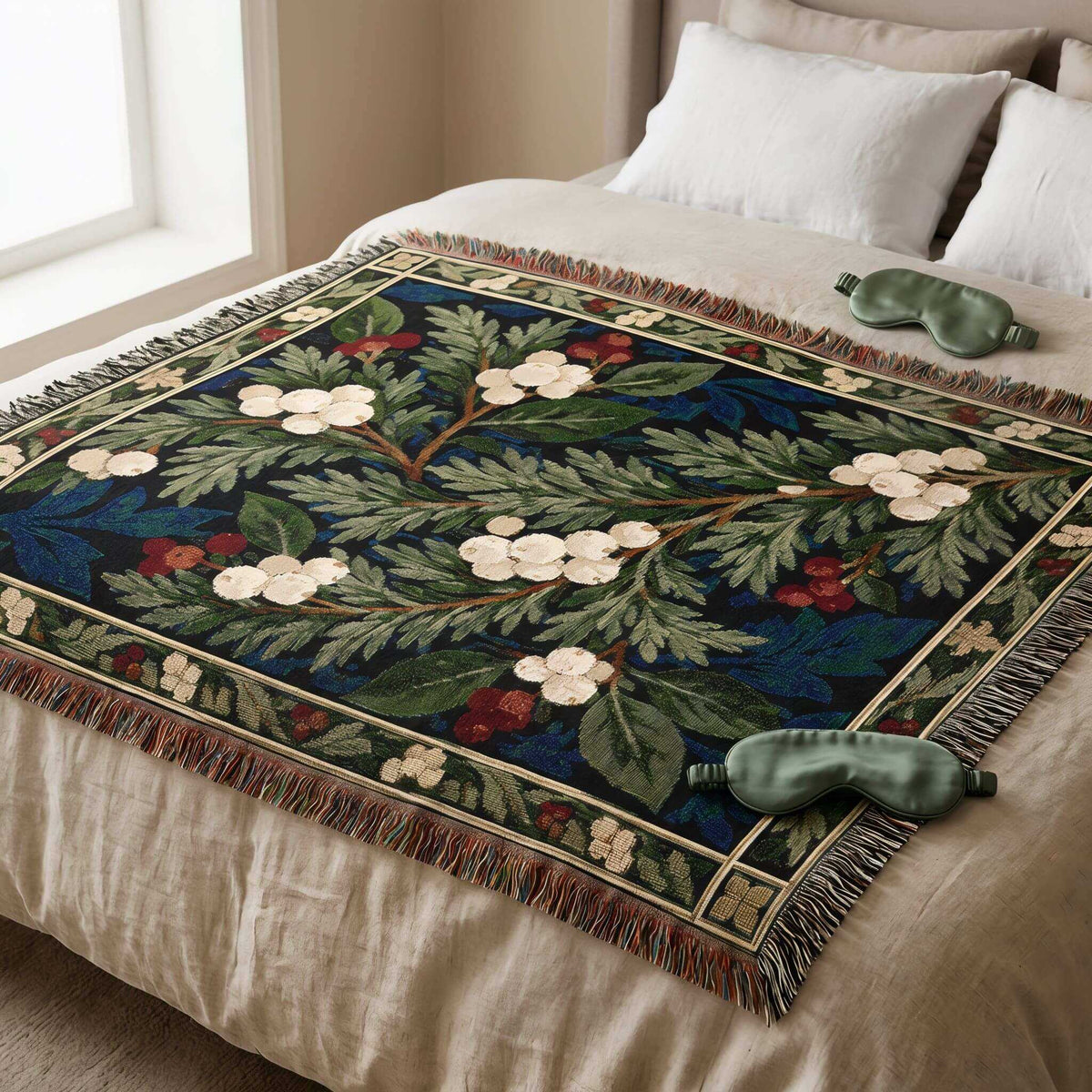 A winter tapestry throw blanket with a floral pattern is draped over a bed. The blanket features a dark navy blue background with green leaves, white flowers, and small red berries, finished with a fringed border.