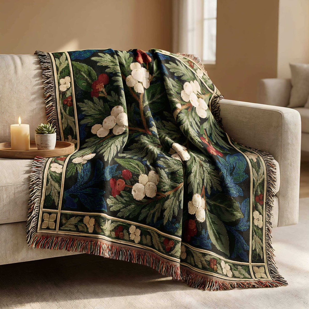 A winter styled woven throw blanket with a floral and leaf pattern draped over a light beige sofa in a living room setting. The blanket features a dark navy blue background with green leaves, white flowers, and red berries, finished with a multicolor fringe.