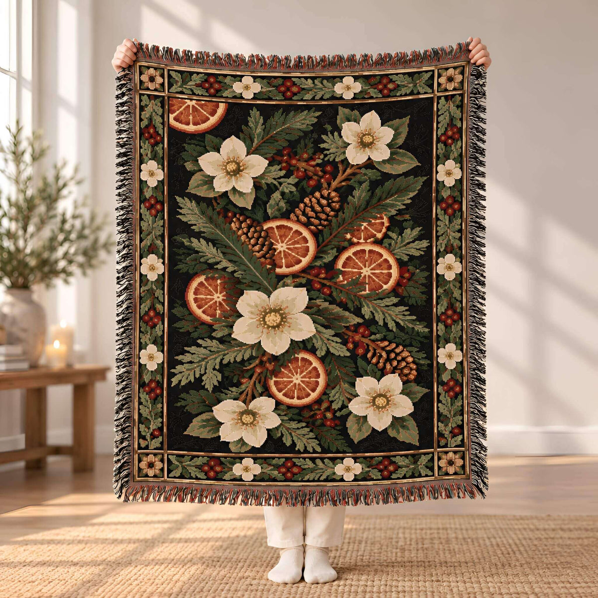 A person holds up a large woven throw blanket with a botanical holiday design featuring white flowers, pinecones, citrus slices, and red berries on a dark background. The blanket has fringe on all sides and is photographed in a bright, modern room with a window and wooden table in the background.