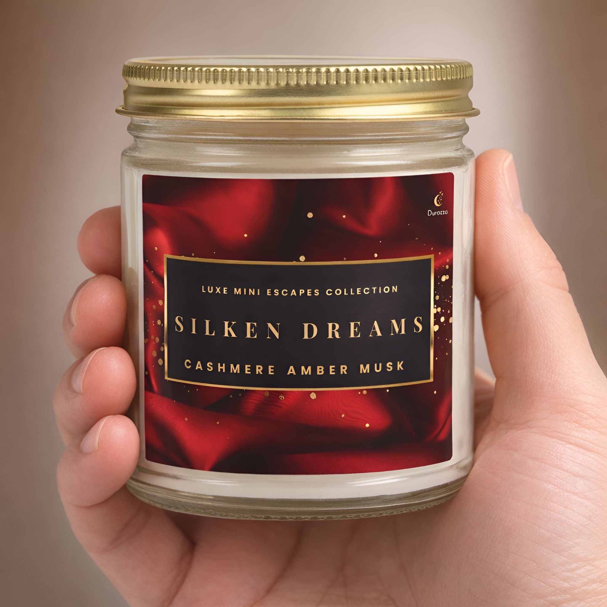 Hand holding a 9oz scented candle jar labeled 'Silken Dreams' with a red and gold design.