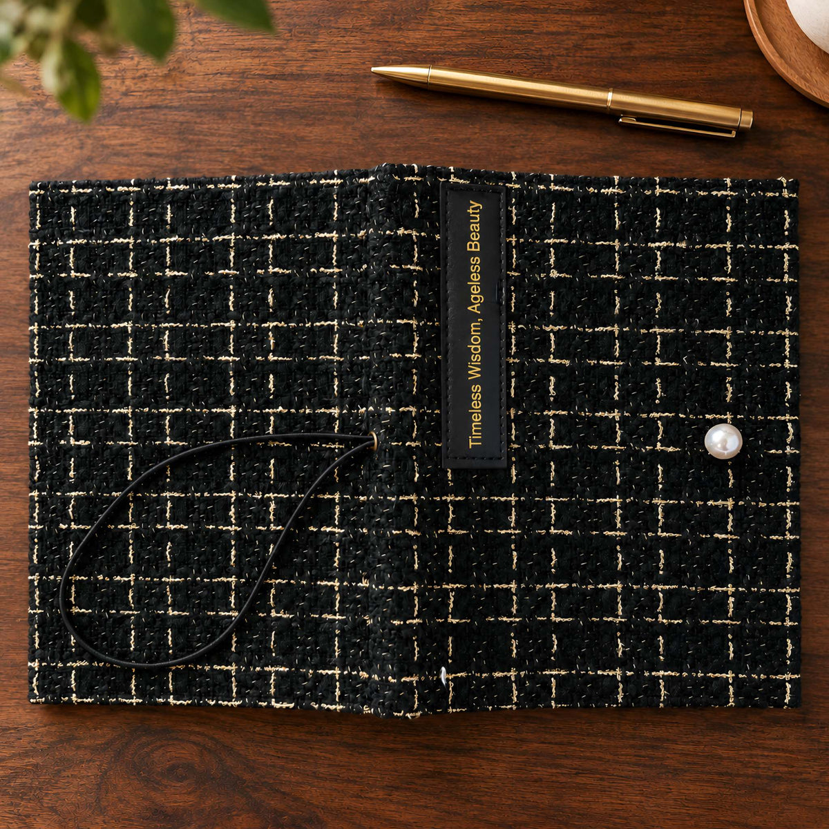 Close-up of black fabric journal cover showing gold-thread weave, Timeless Wisdom label, and pearl-like accent.
