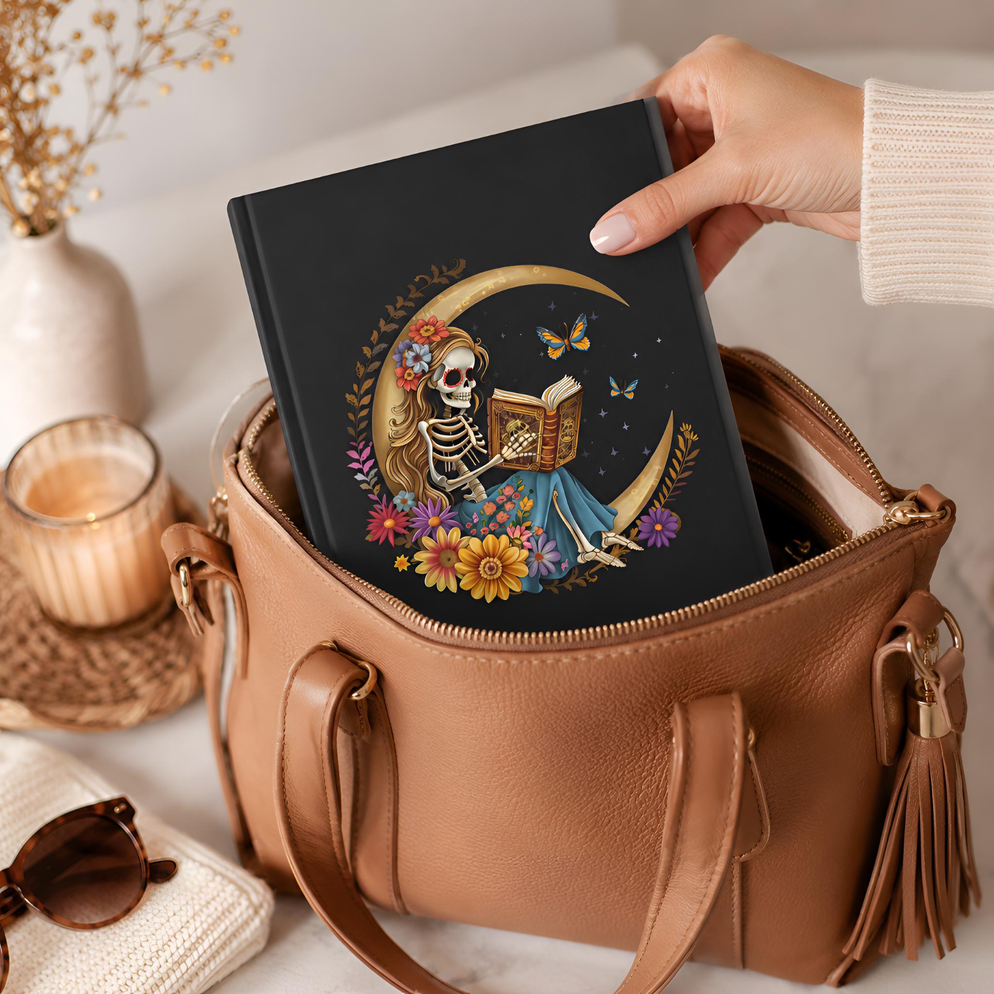 Hand holding a whimsigoth hardcover journal with a skeleton design over a brown leather handbag.