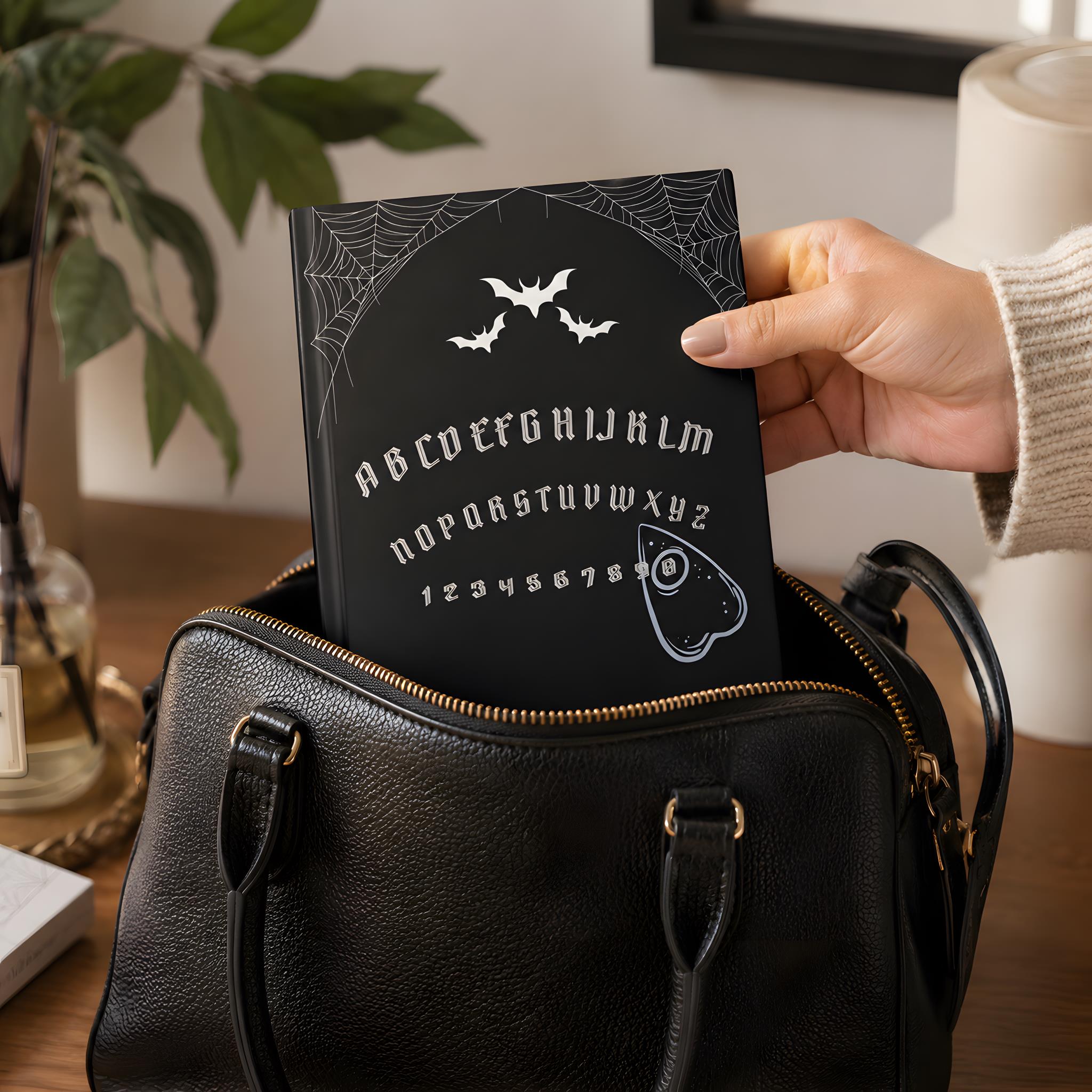 Person holding a ouija board ournal with Halloween-themed design over a black handbag.
