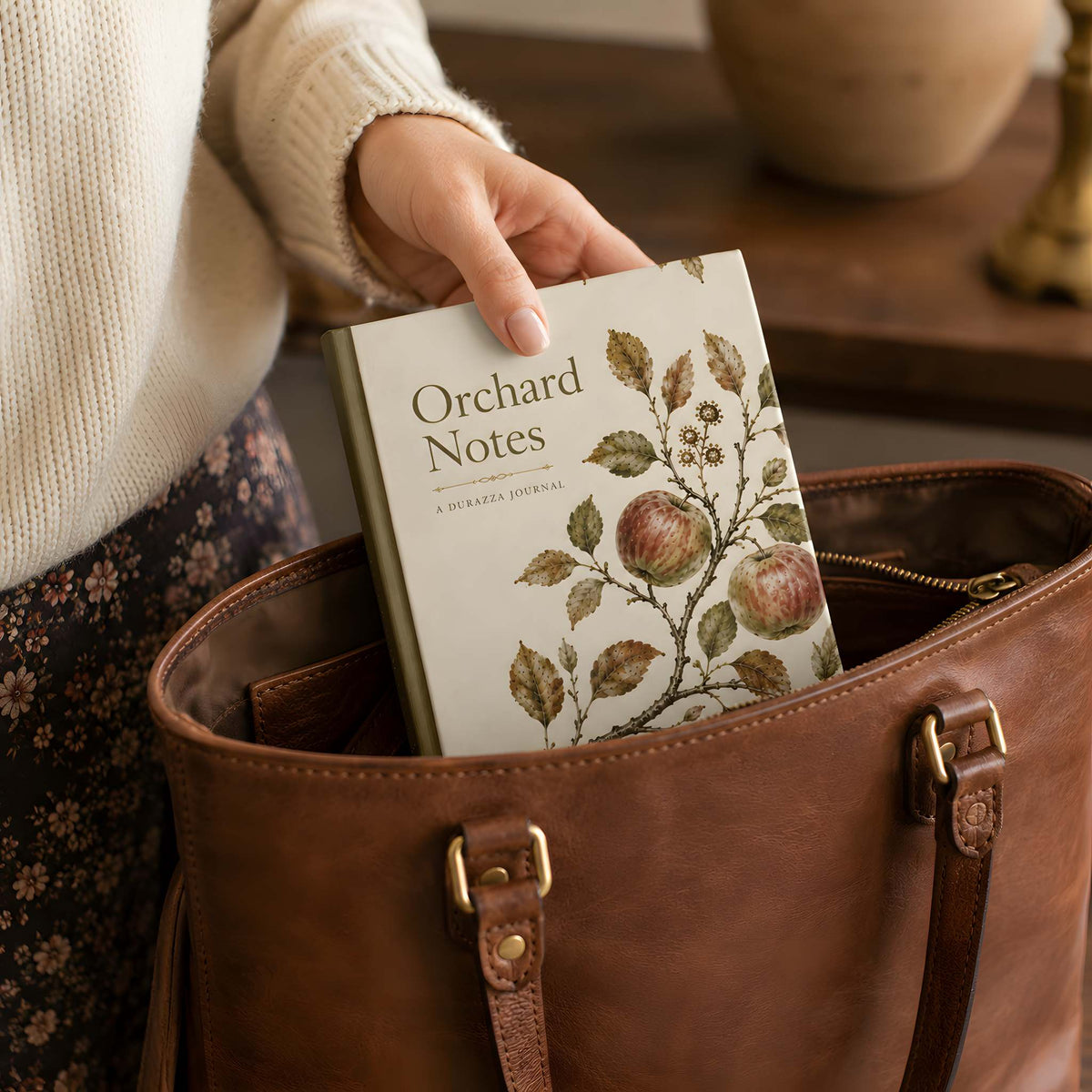 A person in a cream knit sweater and floral skirt places an 'Orchard Notes' journal into a brown leather tote bag.