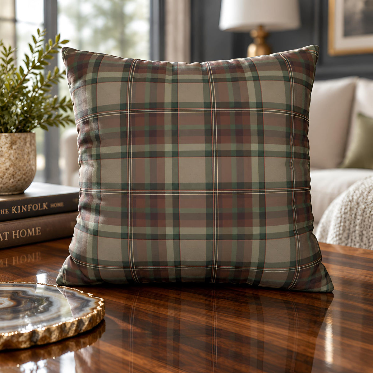A square throw pillow cover with a classic plaid pattern in muted green, beige, and brown tones, styled on a dark wooden table in a cozy living room setting.