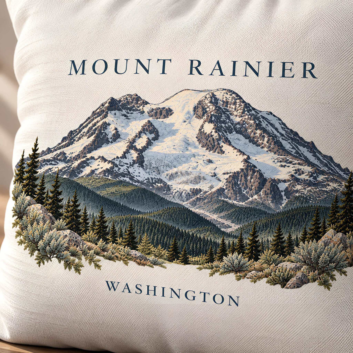 A close-up of a greige-colored throw pillow featuring a detailed embroidered or printed graphic of Mount Rainier in Washington, with the text 'MOUNT RAINIER' at the top and 'WASHINGTON' at the bottom.