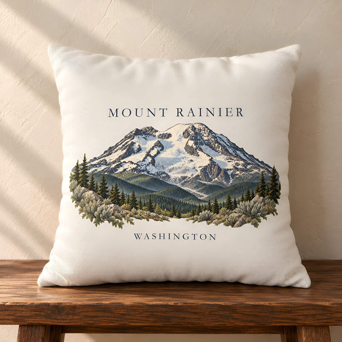 A square off-white throw pillow cover featuring an illustrated graphic of Mount Rainier in Washington, displayed on a rustic wooden bench against a textured beige wall with soft diagonal shadows.