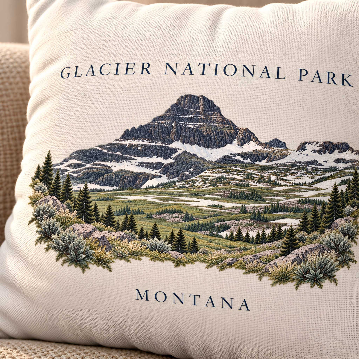 A square off-white throw pillow cover featuring an embroidered scenic design of Glacier National Park, Montana, with a mountain, forests, and meadows, and the text 'GLACIER NATIONAL PARK' and 'MONTANA'.