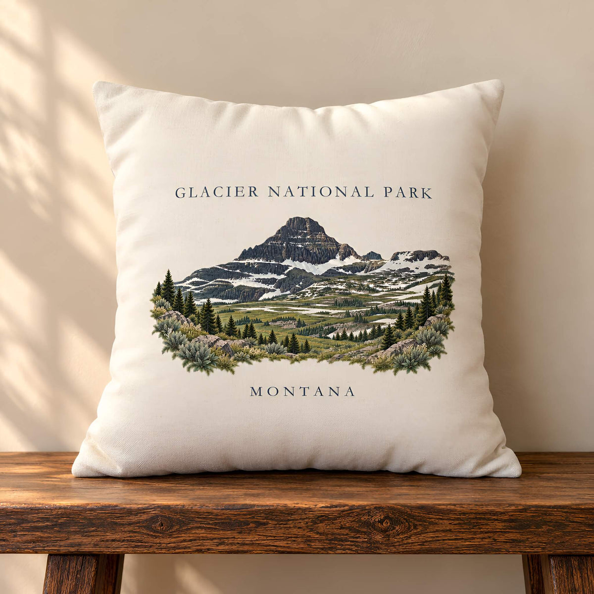 A square cream throw pillow cover featuring an illustrated Glacier National Park mountain scene with the text 'GLACIER NATIONAL PARK' above and 'MONTANA' below, resting on a wooden bench against a beige wall.