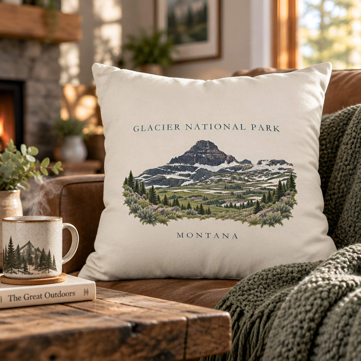 A square ivory throw pillow featuring a scenic illustration of Glacier National Park, Montana, styled on a brown leather couch next to a cozy green knit blanket and a wooden coffee table with a mug.
