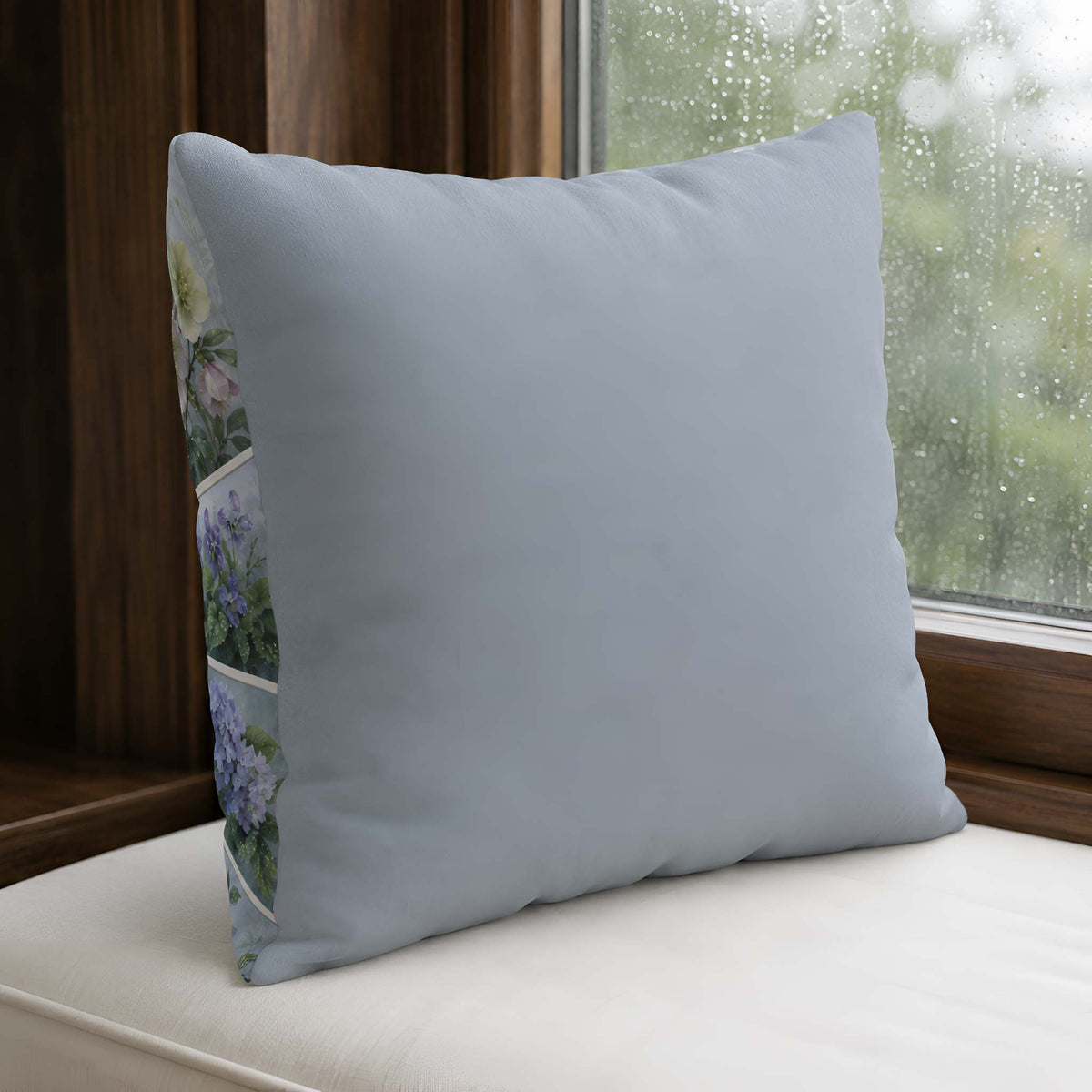 A square blue pillow cover in a dusty light blue-gray color, resting on a cream-colored cushion in front of a window with raindrops. The side of the pillow features a vertical printed panel with a collage of purple hydrangea flowers.