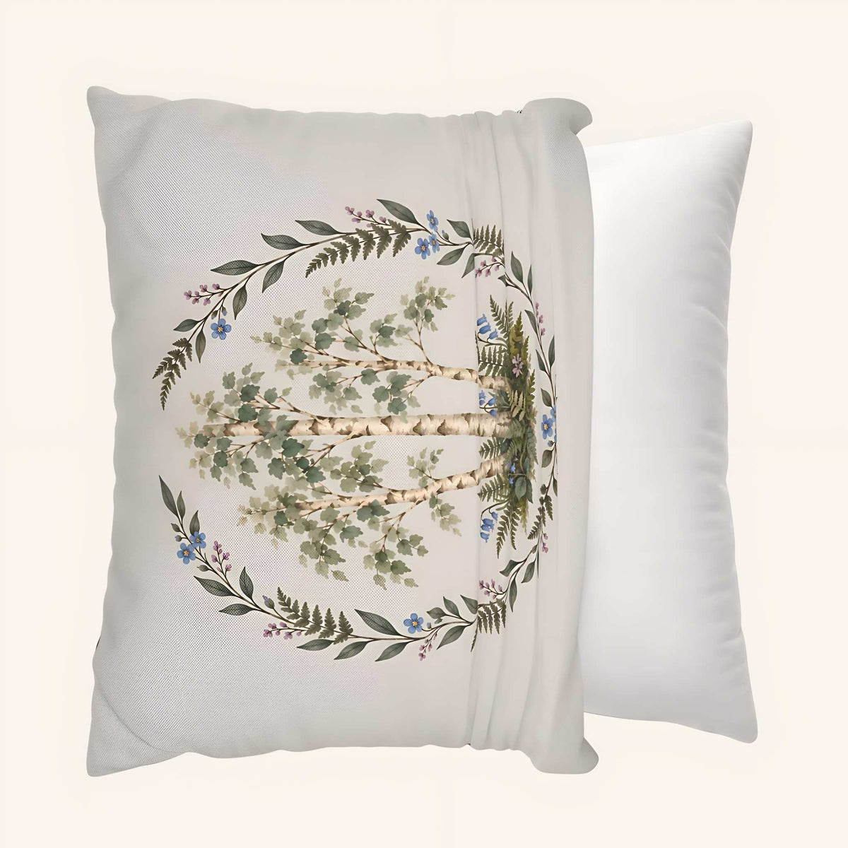A square decorative pillow cover being slipped over a throw pillow. The front pillow has a soft off-white background with a printed botanical design of a tree surrounded by a circular wreath of green leaves and small blue flowers.