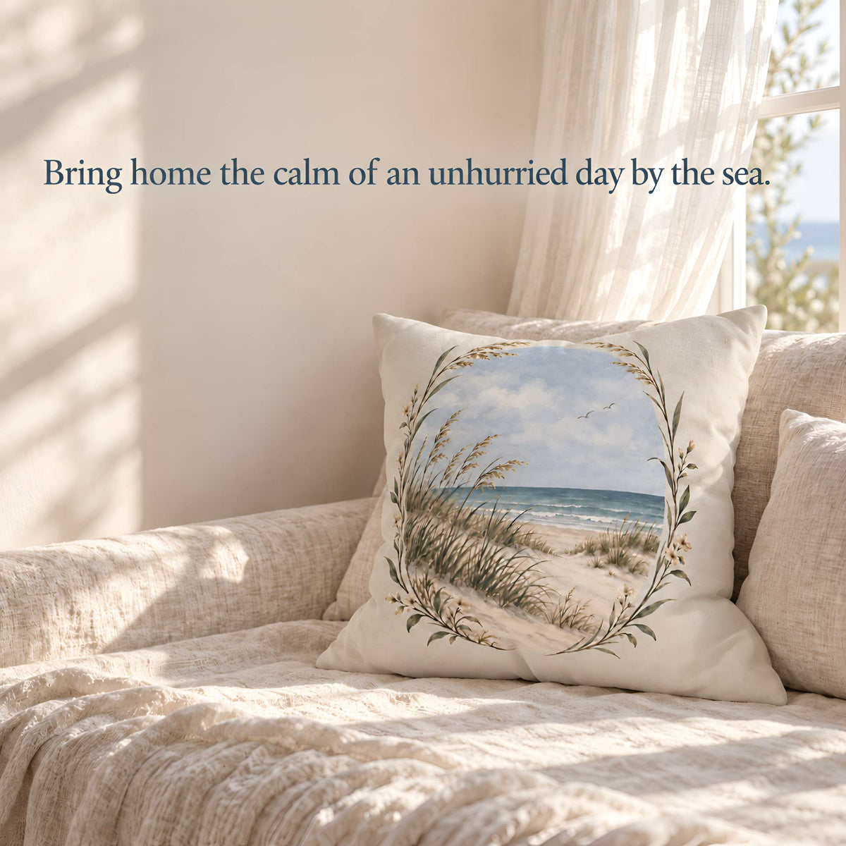 A square decorative throw pillow featuring a beach dunes and ocean scene framed by a leafy wreath, resting on a beige textured sofa in a sunlit room. Text on the left reads, 'Bring home the calm of an unhurried day by the sea.