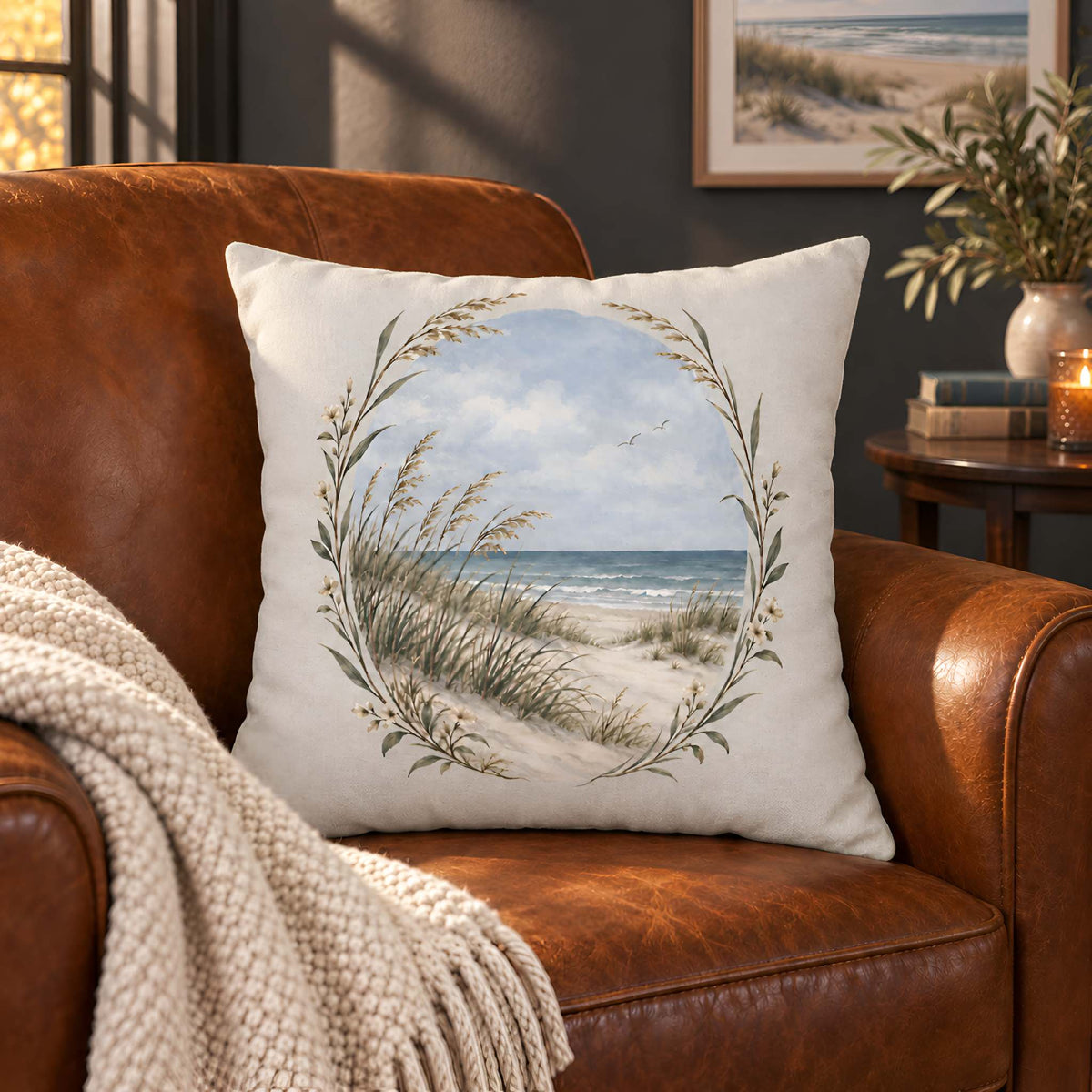 A square throw pillow cover with a beach dune landscape print, styled on a brown leather armchair with a cream knit blanket.