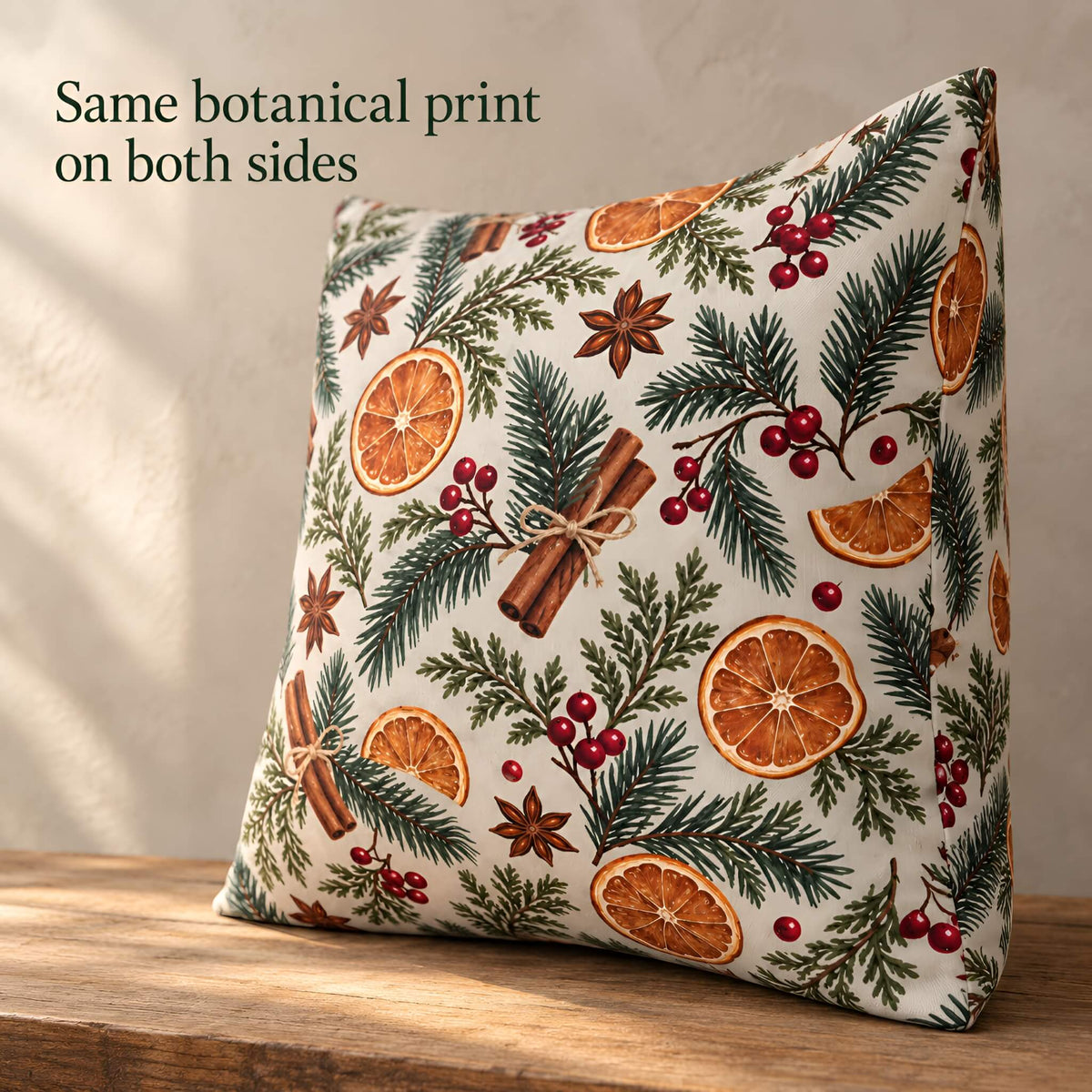 A square decorative pillow cover with a botanical print featuring green pine branches, dried orange slices, red berries, cinnamon sticks, and star anise on an off-white background, displayed on a wooden surface with text overlay 'Same botanical print on both sides'.