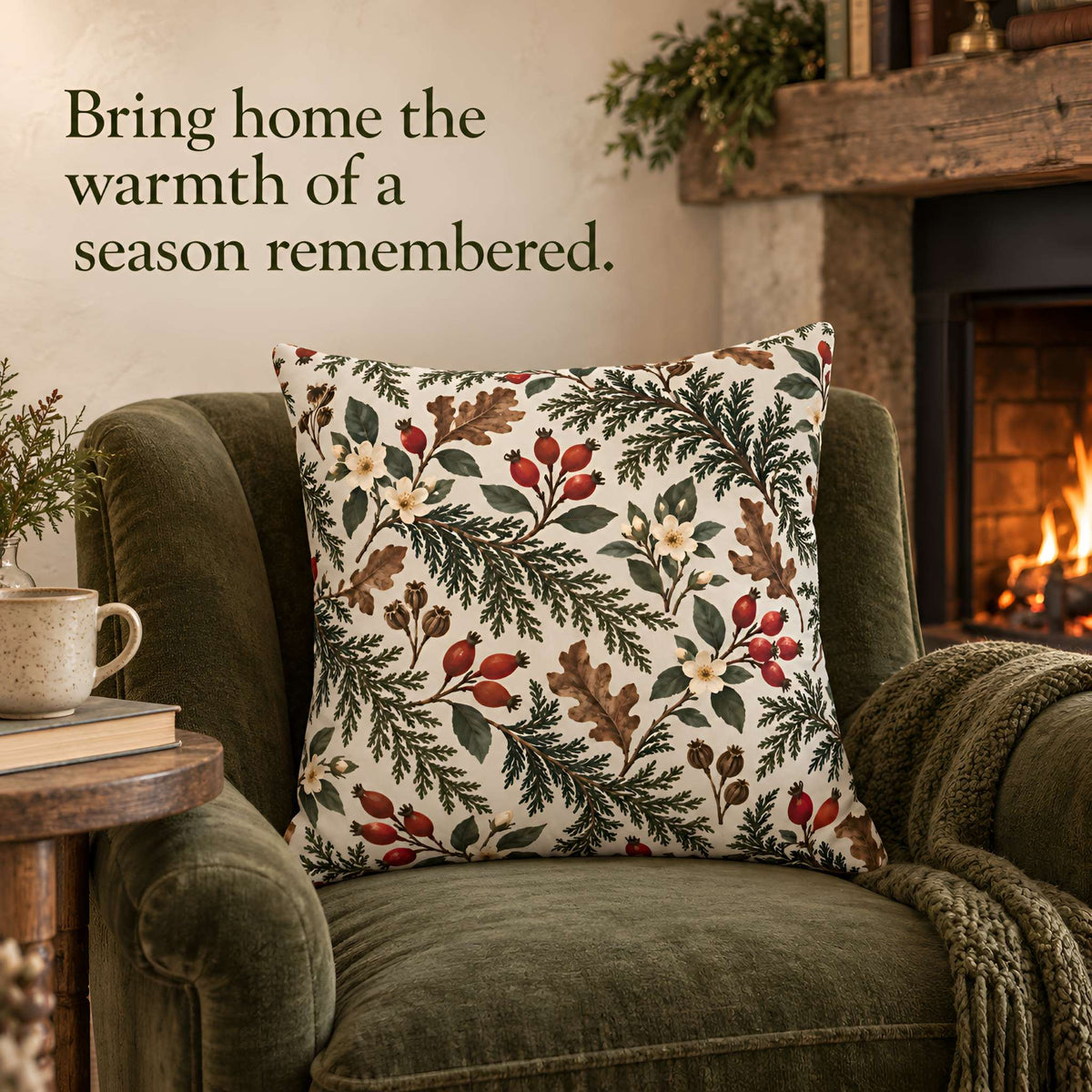 A square decorative throw pillow with a winter botanical pattern of pine branches, oak leaves, and red berries on an off-white background, styled on an olive green armchair in a cozy living room setting with a fireplace and the text 'Bring home the warmth of a season remembered.'.